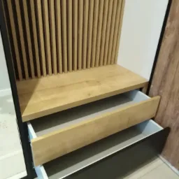 Wnęka z drewnianymi lamelami, blatem i dwiema wysuwanymi szufladami z jasnymi frontami w górnej i ciemnymi w dolnej szufladzie.