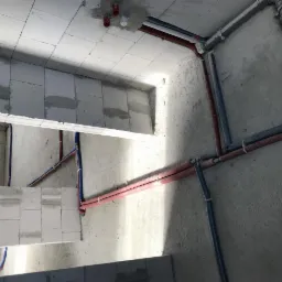 Surowe wnętrze w trakcie budowy z widocznymi rurami instalacyjnymi w kolorach czerwonym i niebieskim, poprowadzonymi po ścianach i suficie z betonu komórkowego.