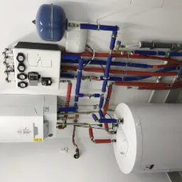 Kocioł grzewczy z zasobnikiem ciepłej wody i rozbudowaną instalacją hydrauliczną z rurami w izolacji czerwonej i niebieskiej, manometry na białej obudowie, widok z dołu.