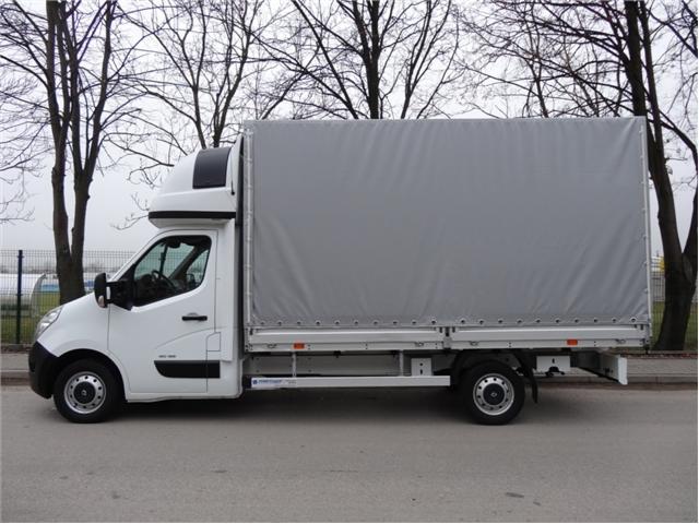 Biały samochód dostawczy z szarą plandeką na tle drzew bez liści i szarej nawierzchni drogi.