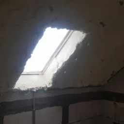 Poddasze z oknem dachowym po aplikacji izolacji. Widoczne nierówności piany i surowy stan ścian z bloczków betonowych.