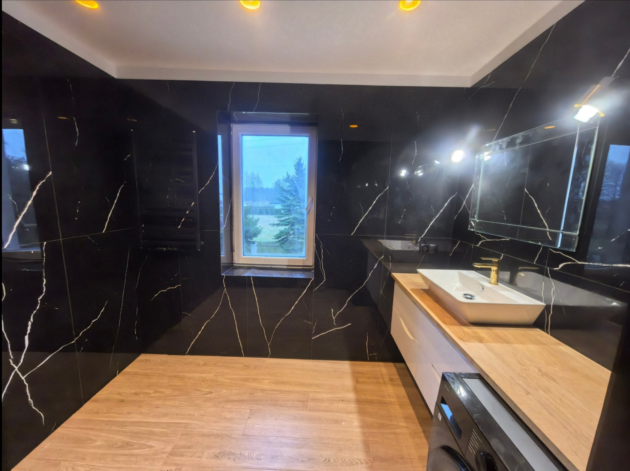 Nowoczesna łazienka z czarnymi płytkami imitującymi marmur, drewnianą podłogą i blatem. Umywalka nablatowa z złotą armaturą. Widok na okno z zielenią na zewnątrz.