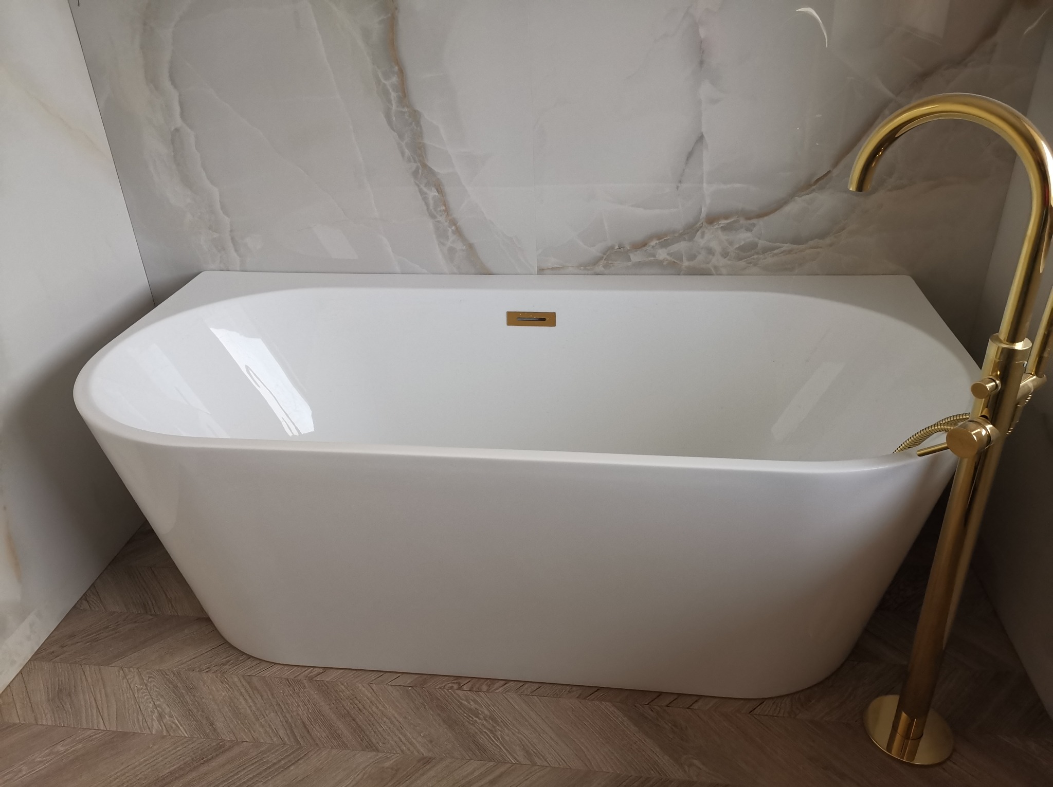 Biała, wolnostojąca wanna z złotą armaturą na tle marmurowych płytek i parkietu ułożonego w jodełkę. Minimalistyczny design łazienki.
