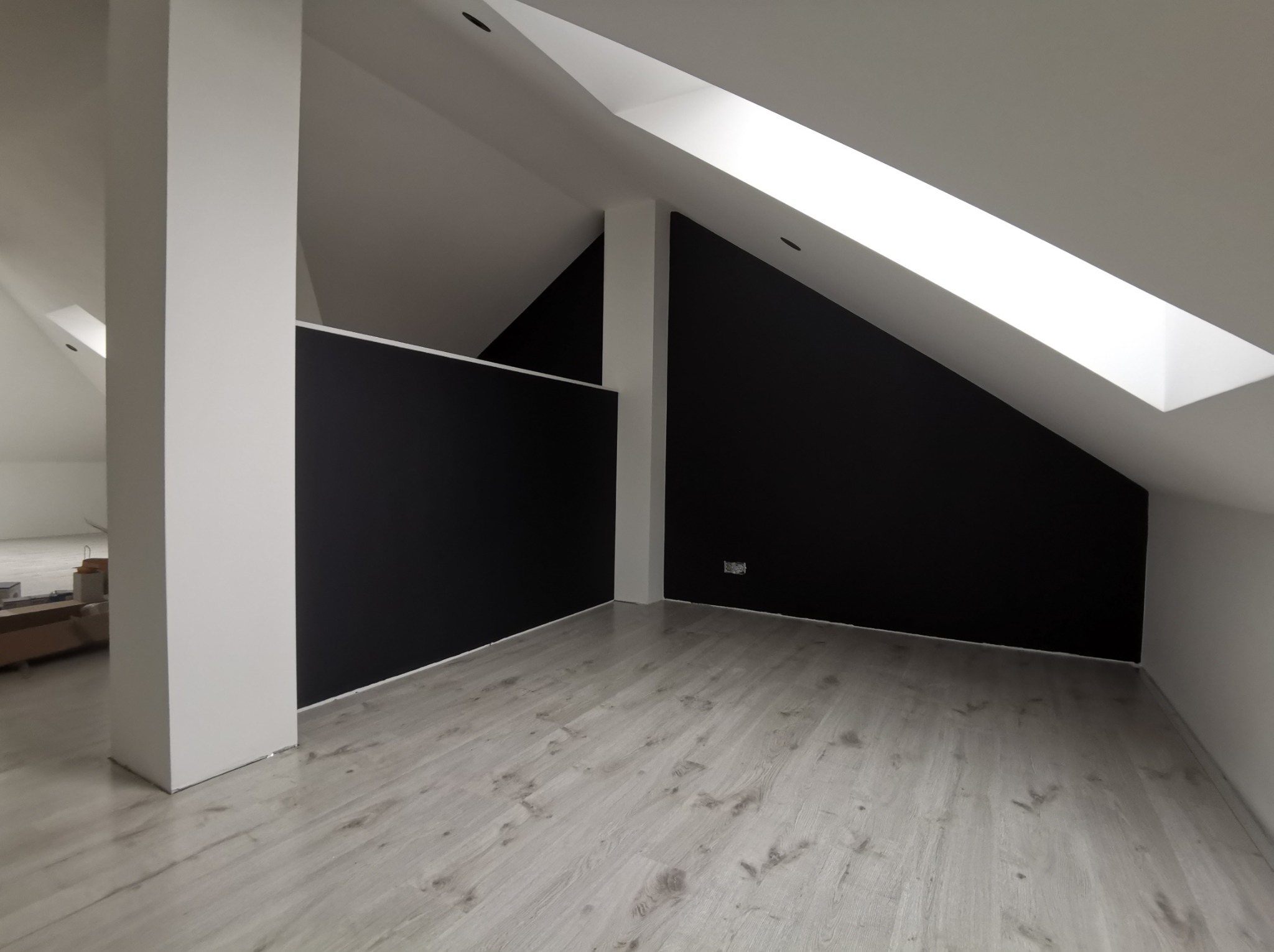 Poddasze po remoncie: czarne ściany, jasna podłoga z paneli, białe elementy architektoniczne i okno dachowe. Minimalistyczny design, nowoczesne wykończenie.