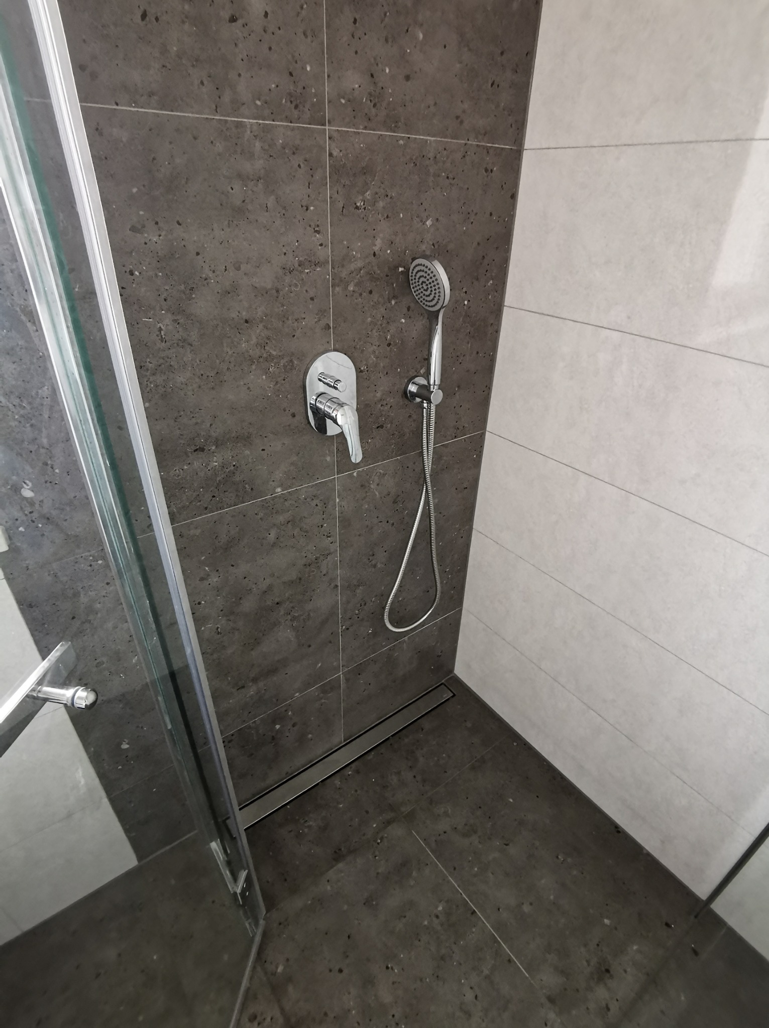 Nowoczesna kabina prysznicowa z ciemnymi płytkami imitującymi kamień i jasnymi, poziomymi płytkami. Widoczna bateria, słuchawka prysznicowa i odpływ liniowy.