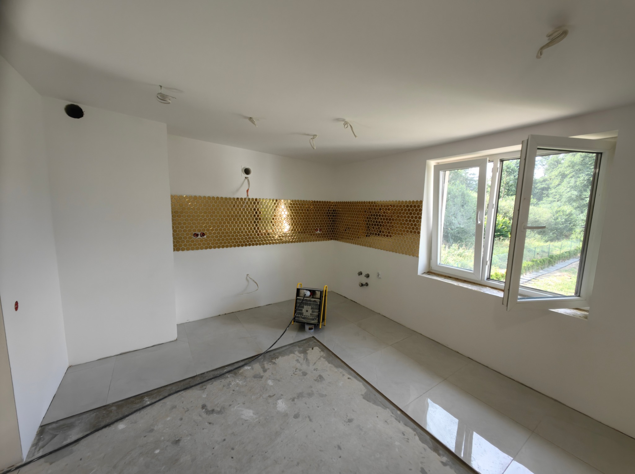 Wnętrze w trakcie wykańczania: złota mozaika na ścianie, częściowo ułożone płytki podłogowe, otwarte okno z widokiem na zewnątrz, nagrzewnica. Etap prac wykończeniowych.