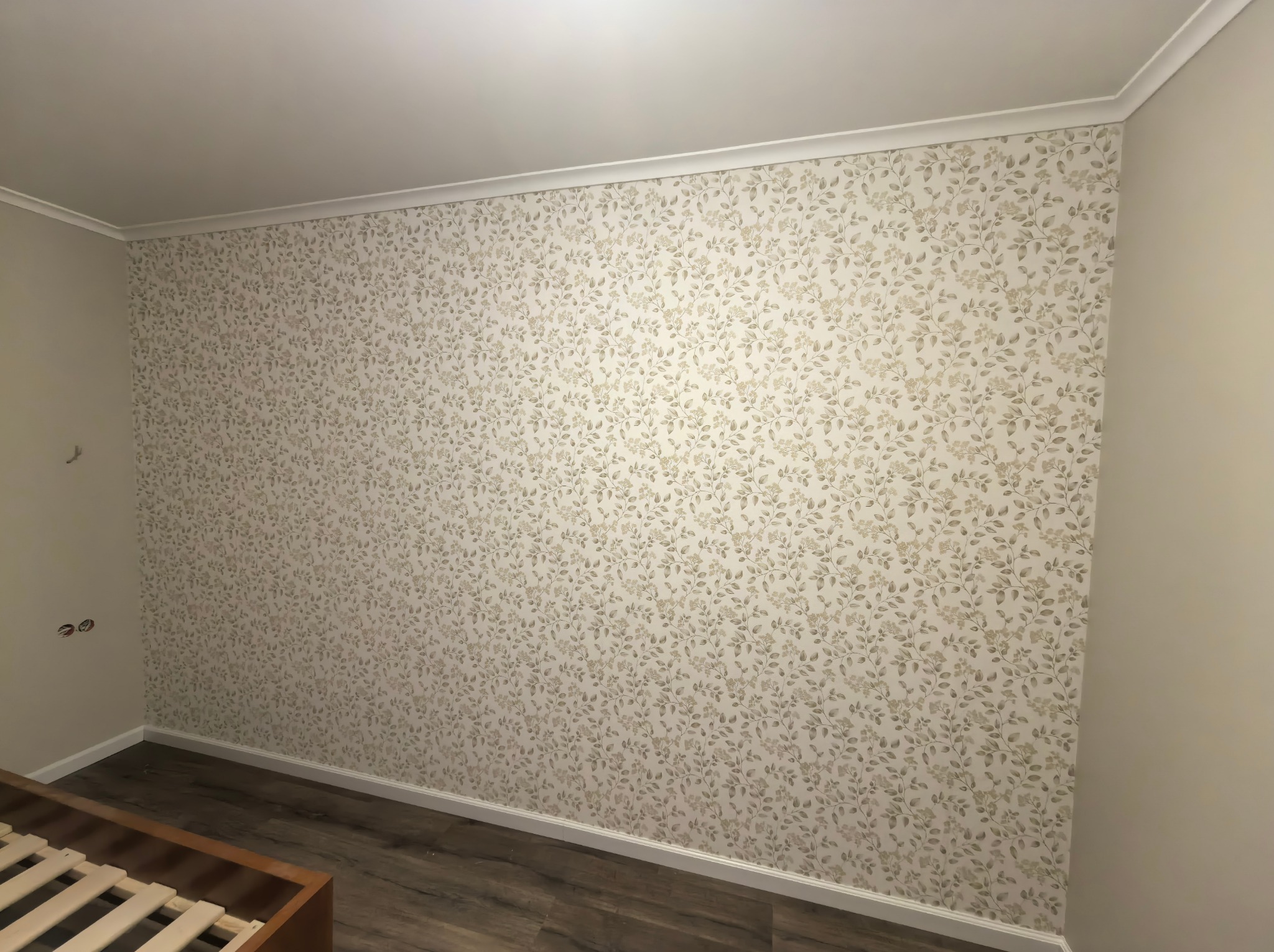 Ściana pokoju wytapetowana w delikatny, roślinny wzór w odcieniach beżu i brązu. Widoczne listwy przypodłogowe i sufitowe. Fragment drewnianego łóżka w dolnym rogu.