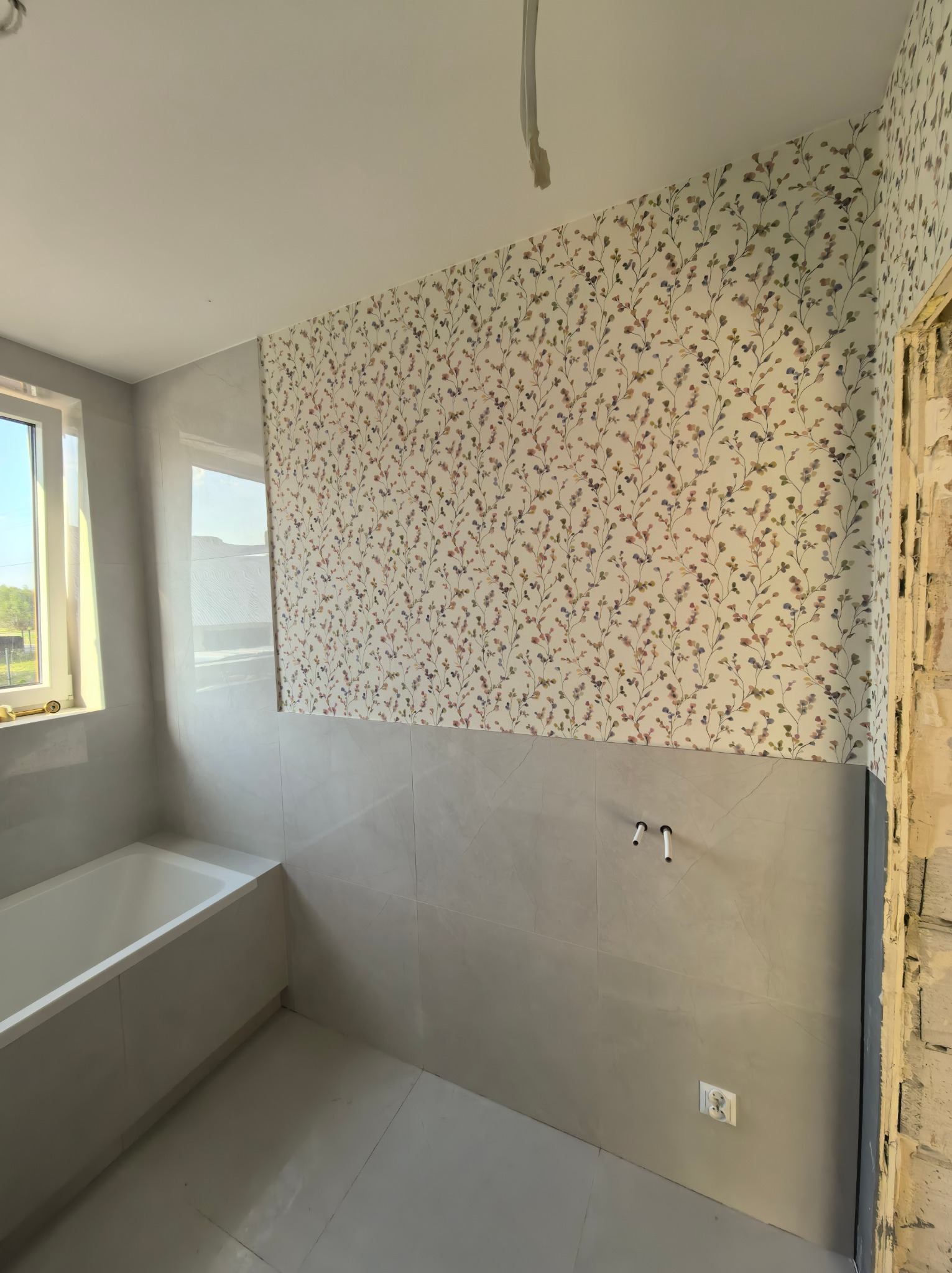 Wykończona łazienka z wanną, szarymi płytkami i tapetą w drobne kwiaty. Widoczne wystające rury i gniazdko elektryczne. Częściowo wykończone obramowanie drzwi.