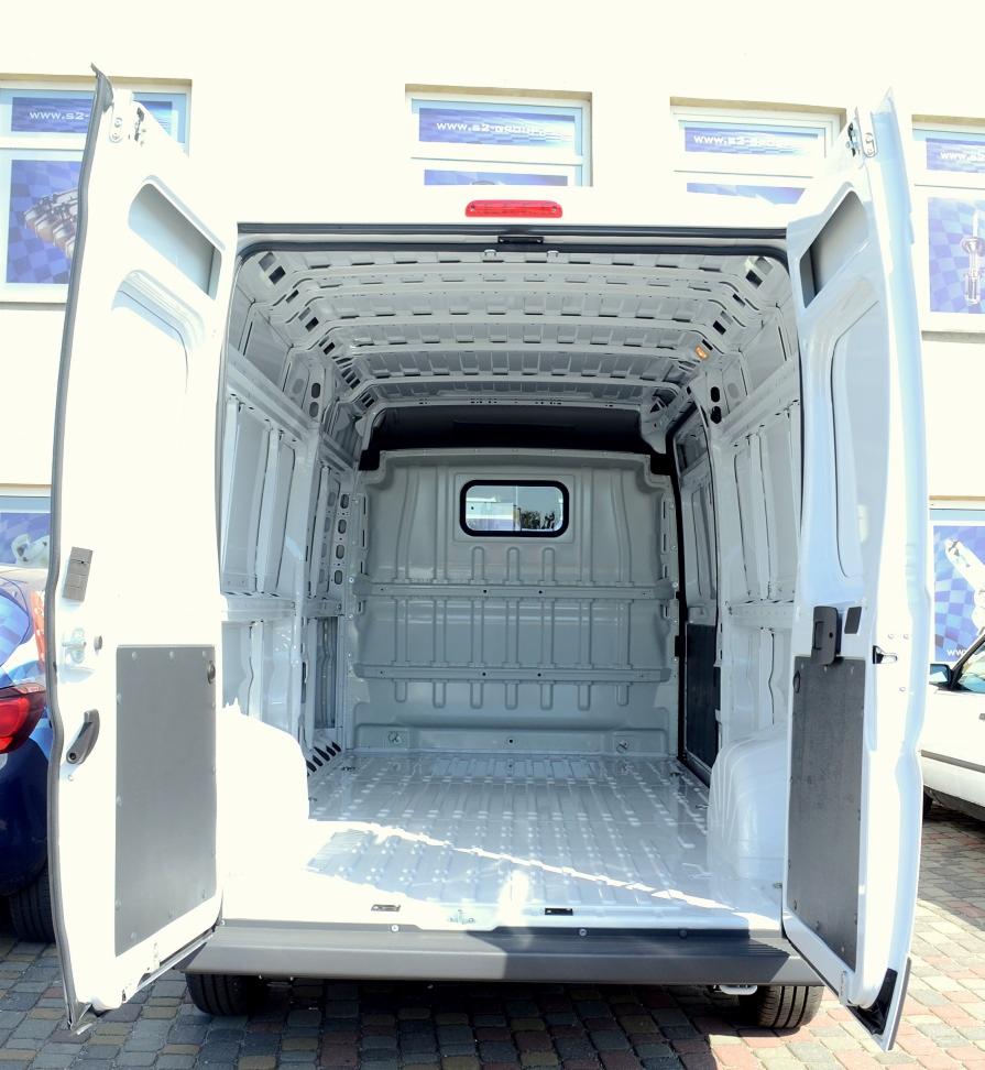 Wnętrze białego, pustego busa z otwartymi drzwiami, widok z tyłu, zarys budynków w tle.