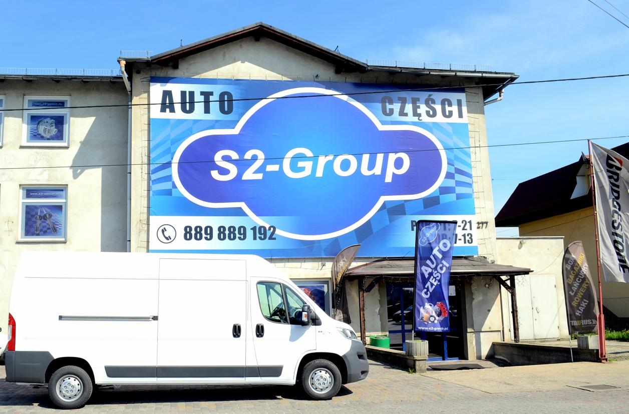 Biały bus dostawczy zaparkowany przed budynkiem z dużym banerem reklamującym sklep z auto częściami S2-Group.