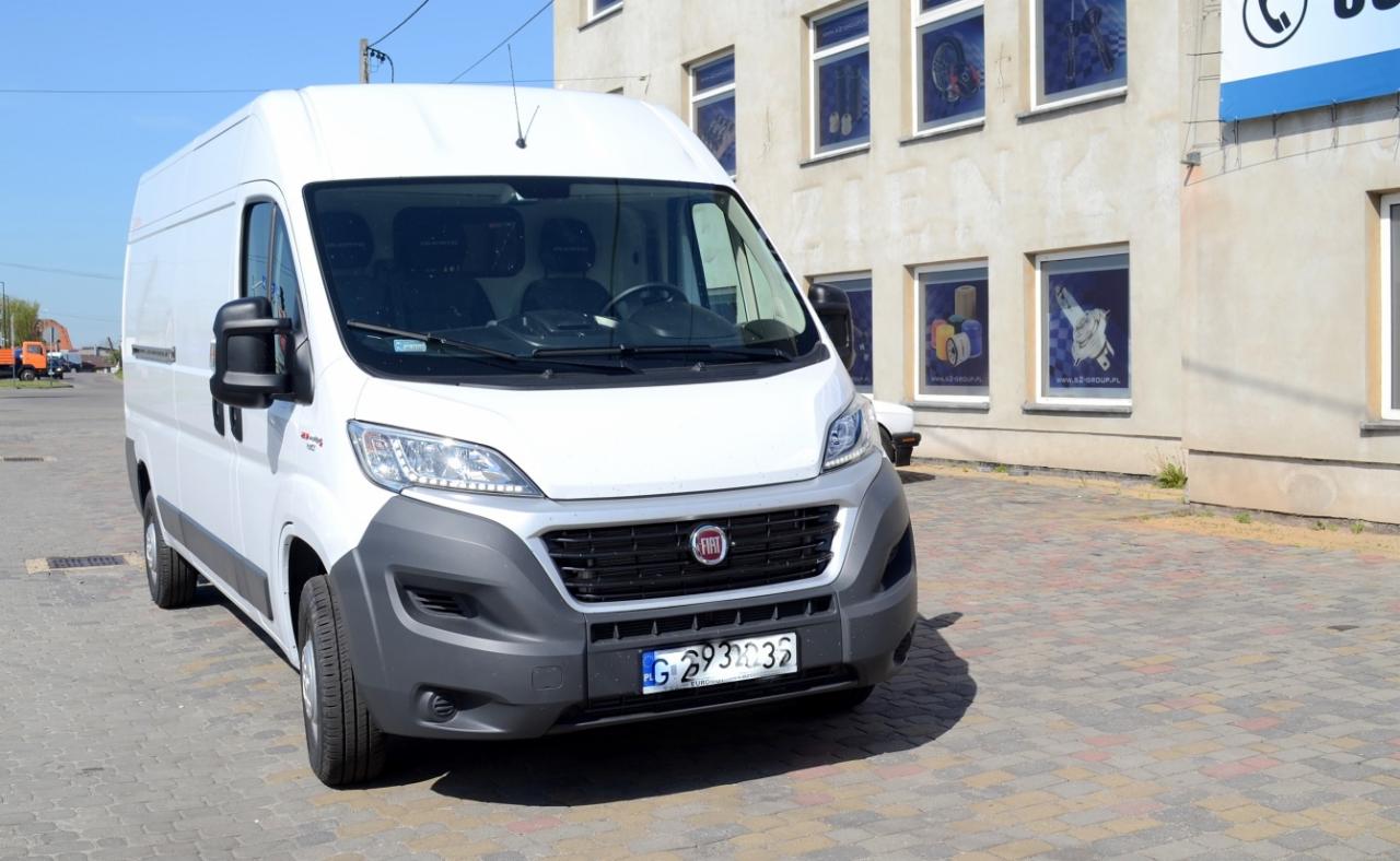 Biały Fiat Ducato, widok z przodu, zaparkowany na brukowanym placu przed budynkiem z oknami ozdobionymi plakatami.