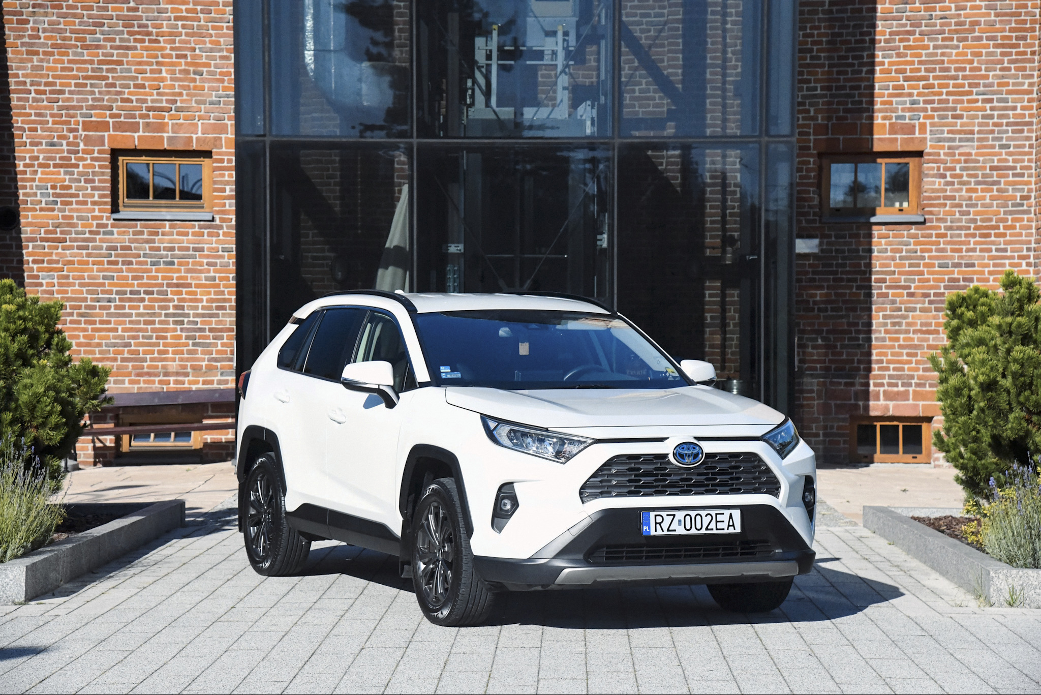 Biały SUV klasy business, Toyota RAV4, zaparkowany przed nowoczesnym budynkiem z cegły i szkła. Widoczne detale: felgi, reflektory, tablica rejestracyjna.