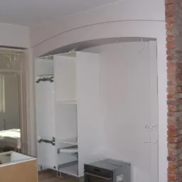 Wnętrze w trakcie remontu: biała szafa bez drzwi z półkami, szary piekarnik elektryczny na podłodze, ściana z czerwonej cegły, przygotowana do malowania ściana z zaznaczonym łukiem i wystającymi...
