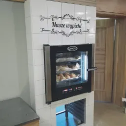 Profesjonalny piec konwekcyjny marki Unox w obudowie z białych płytek, z napisem 'Nasze wypieki' nad nim, prezentujący świeże pieczywo w piekarni.