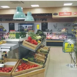 Wnętrze sklepu spożywczego z drewnianymi regałami ekspozycyjnymi wypełnionymi świeżymi warzywami i owocami, oświetlonymi przez wiszące lampy w kolorze miętowym, z widocznymi oznaczeniami...