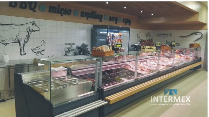 Długa przeszklona lada chłodnicza wypełniona różnymi rodzajami mięs i wędlin, z dekoracyjnymi rysunkami zwierząt na ścianie w tle.