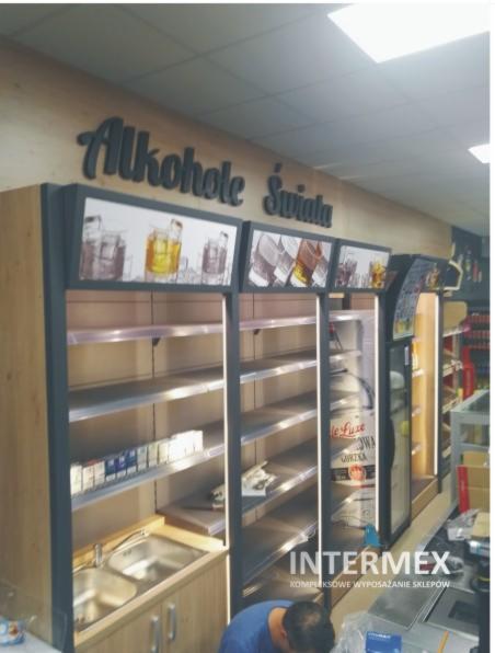 Wnętrze sklepu z regałami na alkohol, podświetlane grafiki napojów, częściowo zaopatrzone w papierosy, z umywalką w dolnej części regału po lewej stronie, widoczny pracownik montujący elementy.