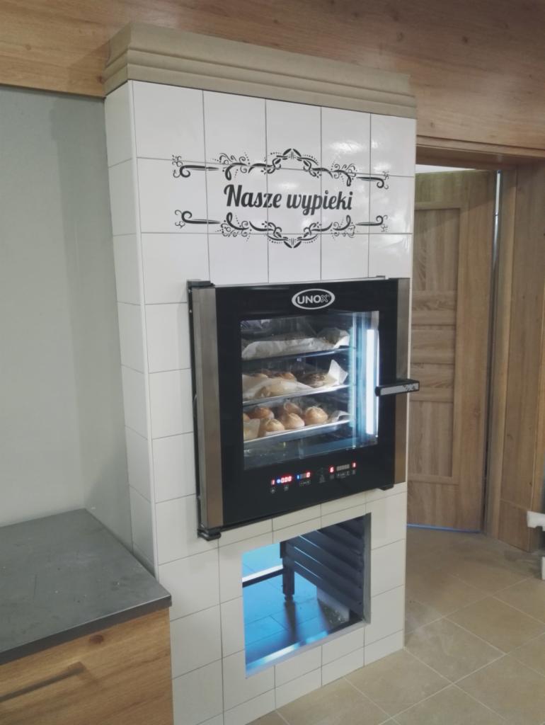 Profesjonalny piec konwekcyjny marki Unox w obudowie z białych płytek, z napisem 'Nasze wypieki' nad nim, prezentujący świeże pieczywo w piekarni.