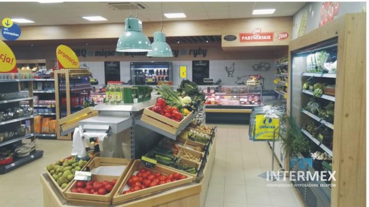 Wnętrze sklepu spożywczego z drewnianymi regałami ekspozycyjnymi wypełnionymi świeżymi warzywami i owocami, oświetlonymi przez wiszące lampy w kolorze miętowym, z widocznymi oznaczeniami...