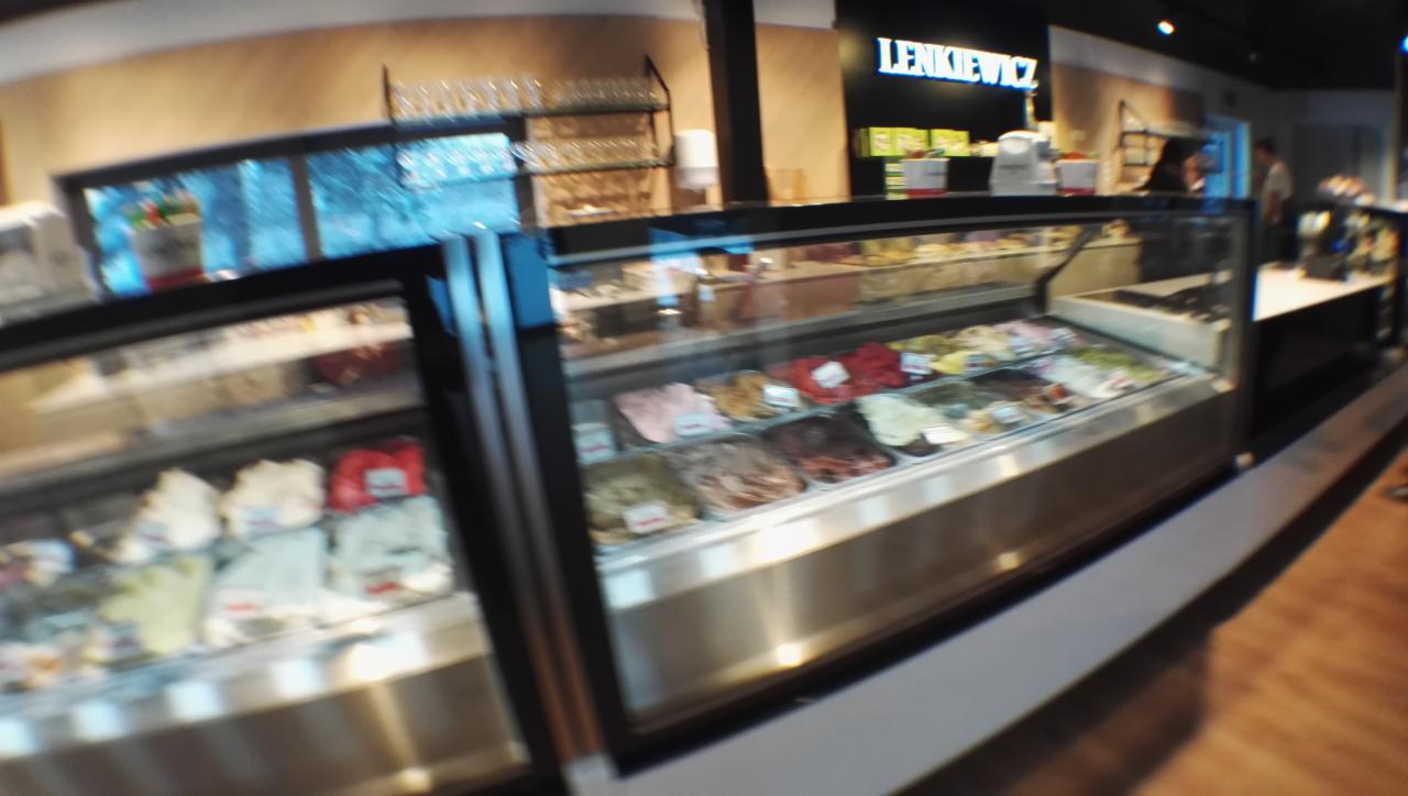 Szeroki kadr na witrynę chłodniczą z różnymi smakami lodów w kawiarni 'Lenkiewicz', widoczne również szklanki na półce i fragment wnętrza lokalu.