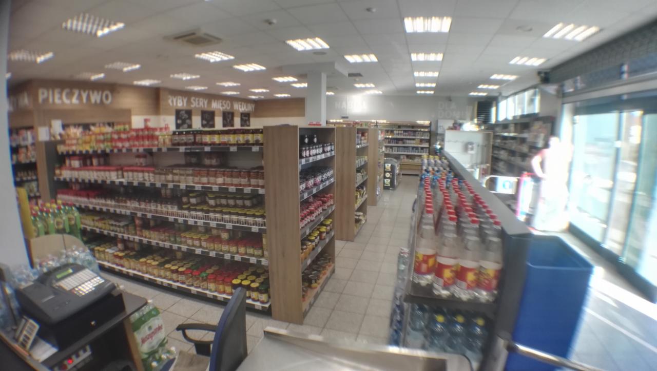 Wnętrze sklepu spożywczego z regałami wypełnionymi produktami, widok z perspektywy kasjera, z widocznym stanowiskiem kasowym i chłodzonymi napojami w pobliżu wyjścia.