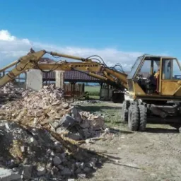 Żółta koparka Zeppelin M13 usuwa gruz po rozbiórce budynku na tle błękitnego nieba i zieleni.