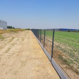 Montujemyogrodzenia.pl - Długa siatka ogrodzeniowa z podmurówką betonową oddzielająca teren piaszczysty od zielonego pola, z budynkami magazynowymi w tle pod błękitnym niebem.