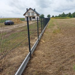Montujemyogrodzenia.pl - Długa linia czarnego ogrodzenia panelowego z betonową podmurówką, oddzielająca pole od posesji z budynkiem w trakcie budowy i zaparkowanym czarnym samochodem.