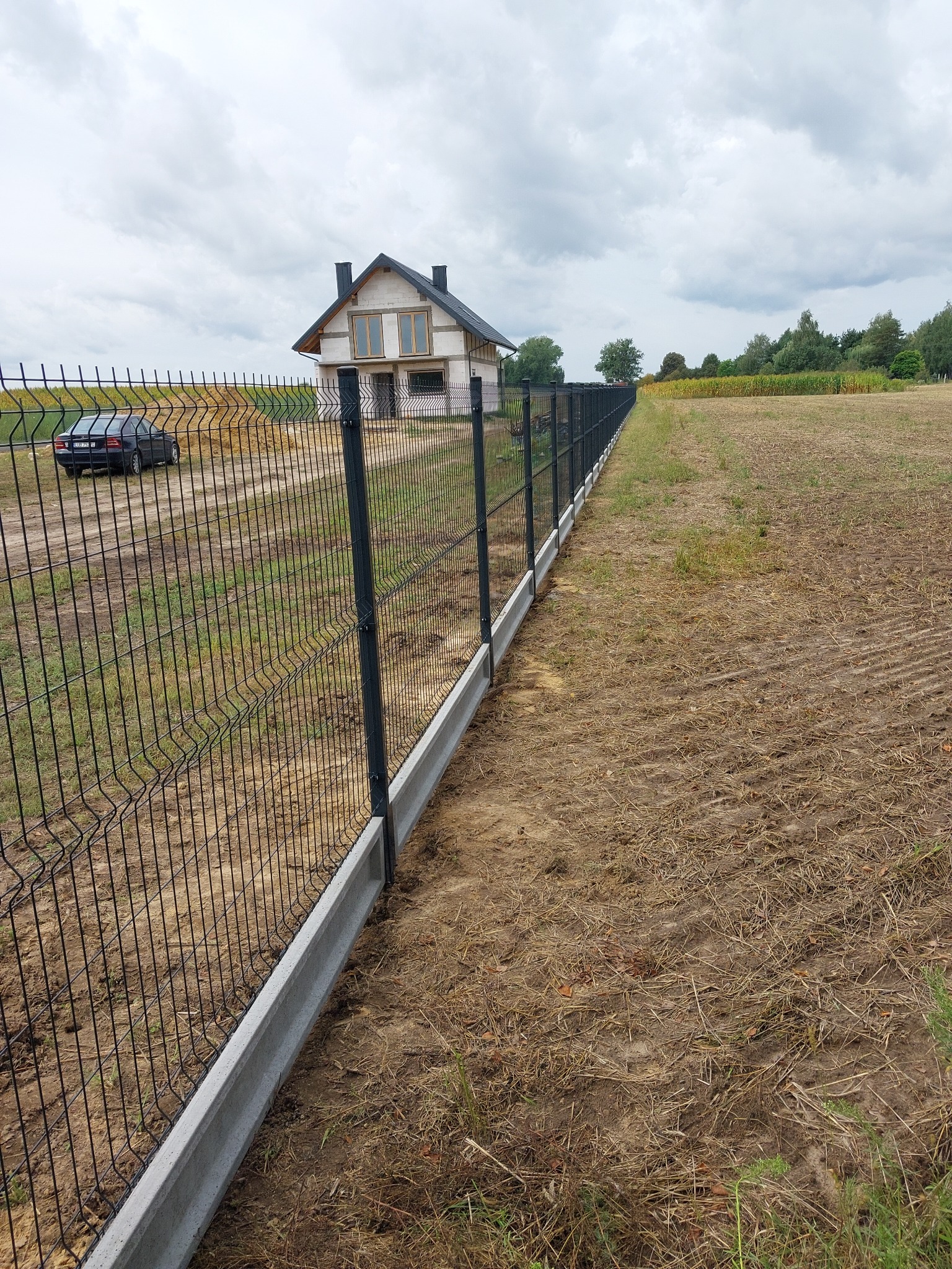 Długa linia czarnego ogrodzenia panelowego z betonową podmurówką, oddzielająca pole od posesji z budynkiem w trakcie budowy i zaparkowanym czarnym samochodem.