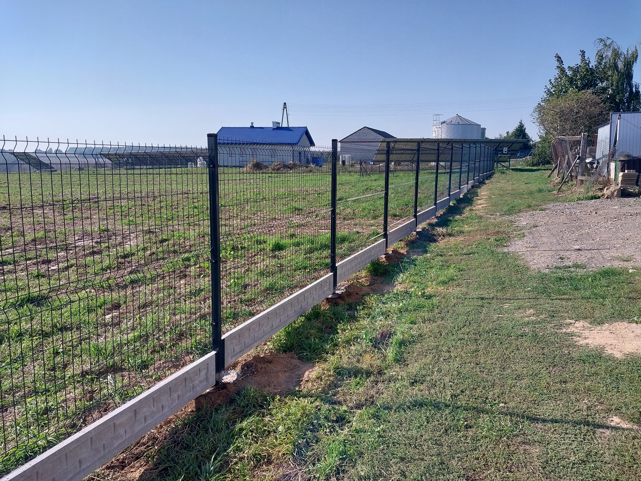 Długi fragment ogrodzenia z siatki z podmurówką imitującą cegłę, widok na pole i zabudowania gospodarcze w tle.