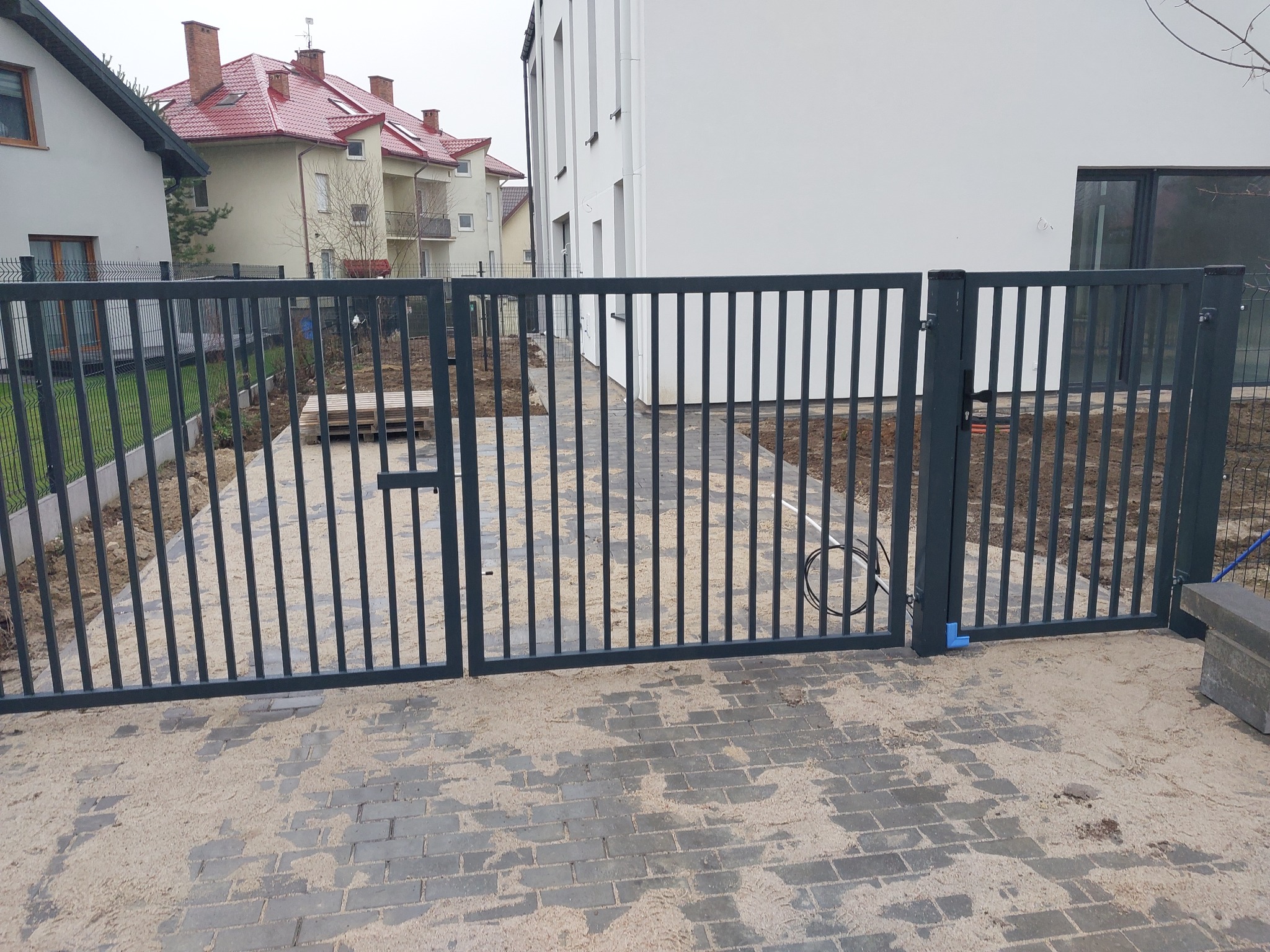 Nowoczesna, grafitowa brama dwuskrzydłowa z pionowych prętów, zamontowana na posesji z brukowaną ścieżką i częściowo zagospodarowanym terenem.