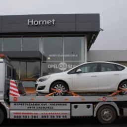 Laweta z białym Oplem Astrą zabezpieczonym pomarańczowymi pasami transportowymi na tle salonu samochodowego Opel Hornet.