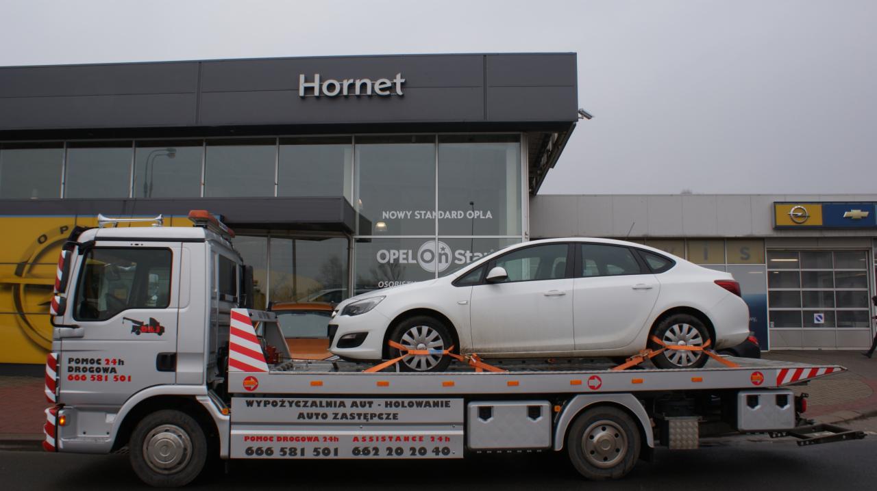 Laweta z białym Oplem Astrą zabezpieczonym pomarańczowymi pasami transportowymi na tle salonu samochodowego Opel Hornet.