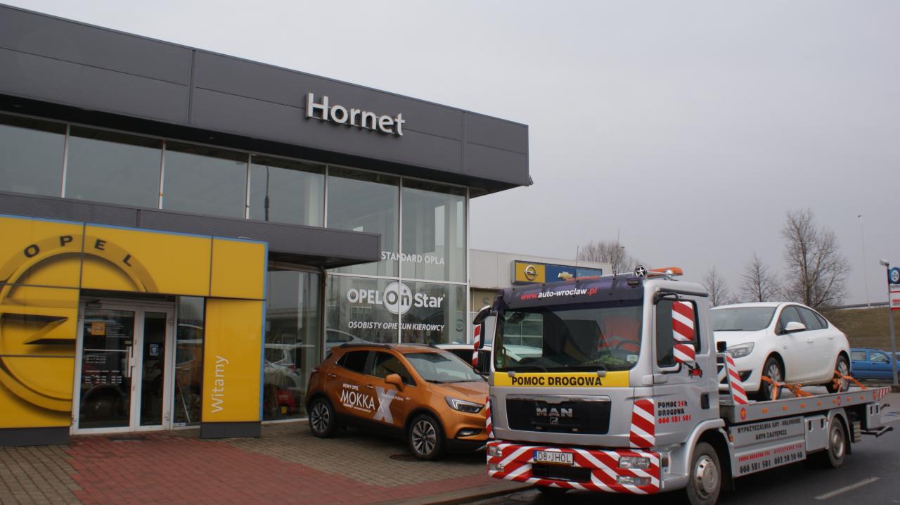 Laweta z białym samochodem na platformie przed salonem samochodowym Opel Hornet we Wrocławiu, widoczny pomarańczowy Opel Mokka.