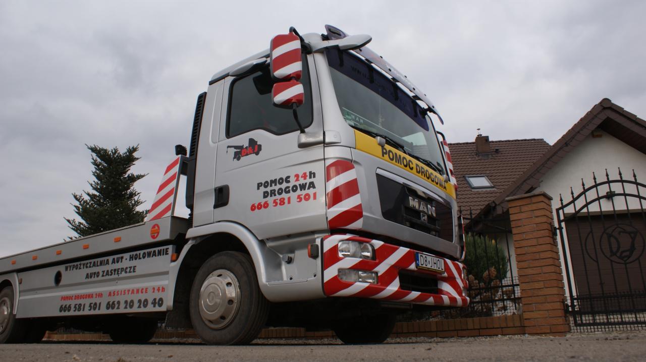 Srebrna laweta MAN z czerwono-białymi pasami i napisem 'Pomoc Drogowa 24h' zaparkowana przed ogrodzeniem domu z brązowym dachem w pochmurny dzień.