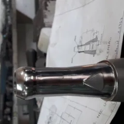 Błyszczący, metalowy element z trójkątnym otworem, leżący na rysunku technicznym z odręcznymi notatkami, w tle widoczny fragment obrabiarki.