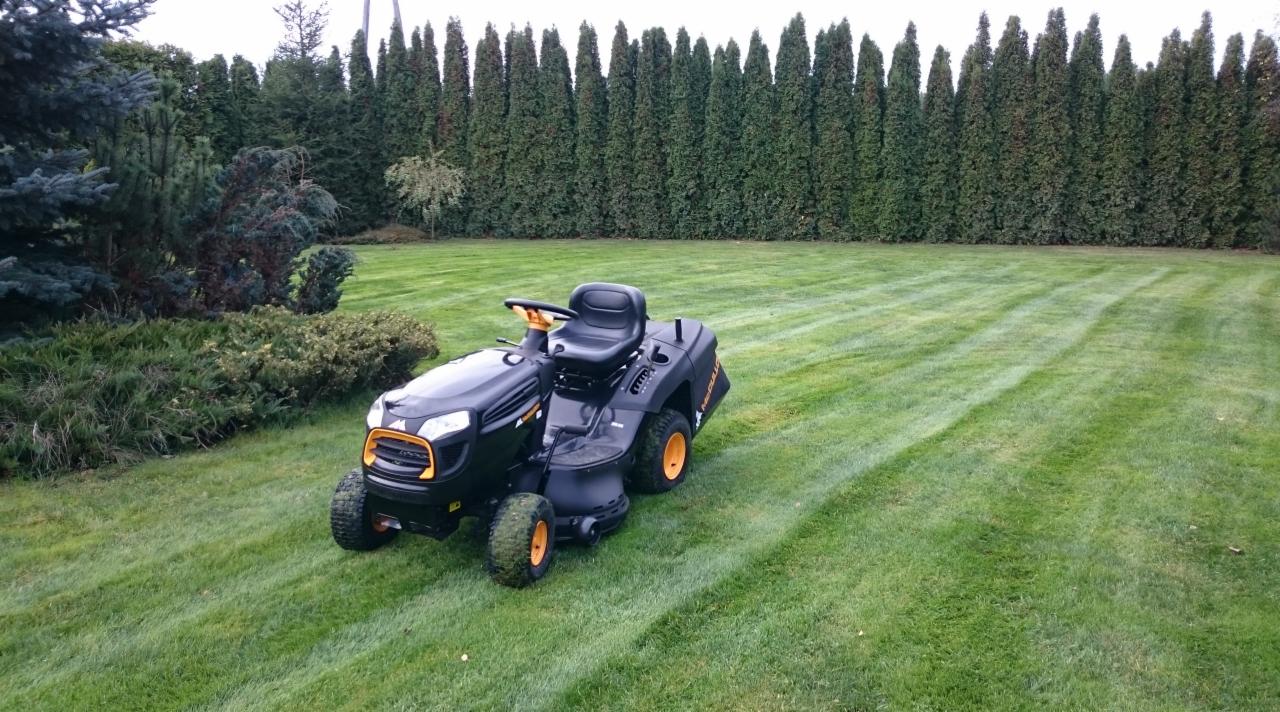 Czarny traktorek do koszenia trawy z żółtymi akcentami na tle równo przystrzyżonego trawnika z widocznymi pasami po koszeniu i szpalerem wysokich drzew.