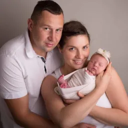 Portret rodzinny: uśmiechnięta mama trzyma zawinięte niemowlę z ozdobną opaską, obok stoi tata w białej koszulce polo na tle szarego tła.