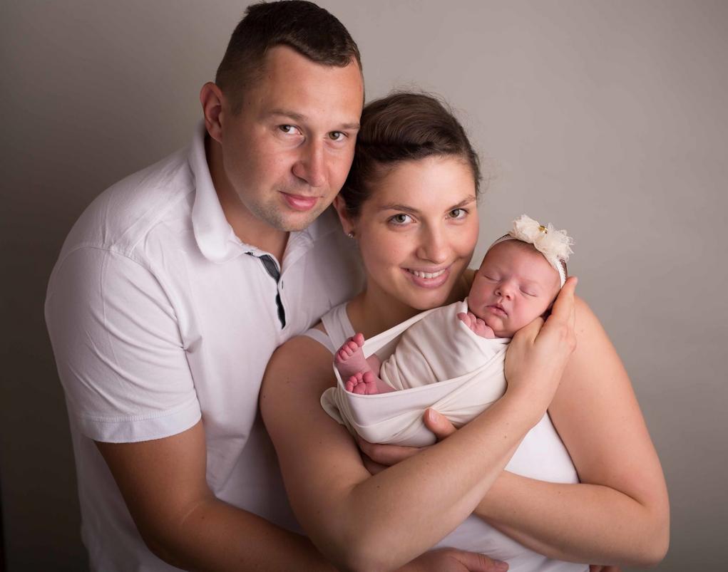 Portret rodzinny: uśmiechnięta mama trzyma zawinięte niemowlę z ozdobną opaską, obok stoi tata w białej koszulce polo na tle szarego tła.