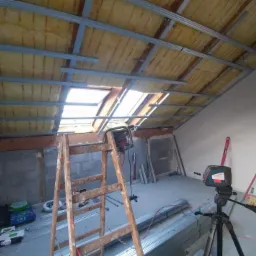 Montaż metalowej konstrukcji pod płyty gipsowo-kartonowe na poddaszu, widoczna izolacja wełną mineralną, laser krzyżowy na statywie i drabina drewniana.