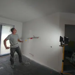 Mężczyzna w szarej koszulce i jeansach wygładza ścianę długą szpachlą na teleskopowym kiju w pokoju z oknem zabezpieczonym folią, widoczne kable elektryczne i lampa budowlana, w tle schody.