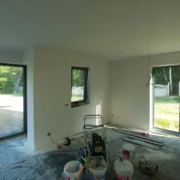 Pokój po świeżym malowaniu, z widocznym agregatem malarskim, wiadrami farby i zabezpieczoną folią podłogą, duże okna z widokiem na zieleń.