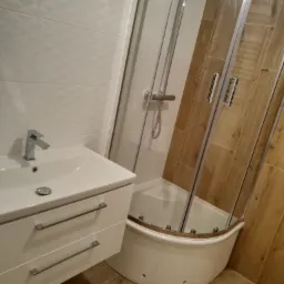 Nowoczesna łazienka z kabiną prysznicową narożną, umywalką z szafką i armaturą w chromowanym wykończeniu, wykończona płytkami w odcieniach bieli i drewna.