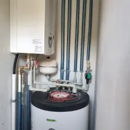 Biały piec gazowy kondensacyjny zamontowany na ścianie, połączony z zasobnikiem ciepłej wody i instalacją z rur w izolacji, widoczne zawory i manometr.