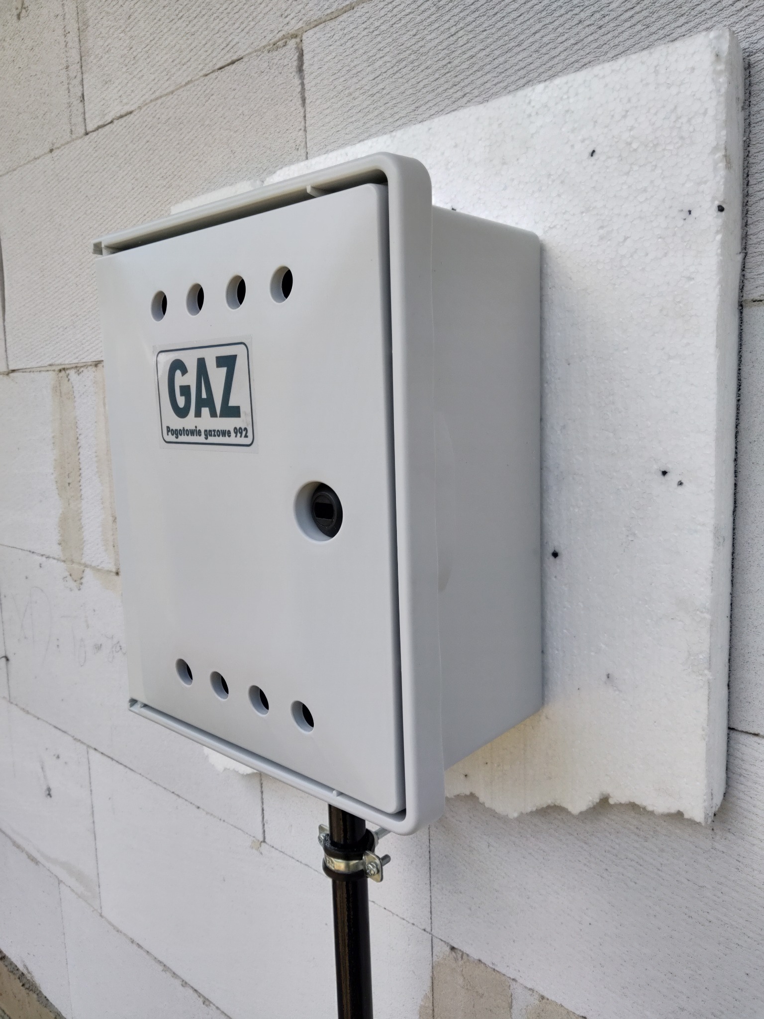 Szara skrzynka gazowa z naklejką 'GAZ Pogotowie gazowe 992' zamontowana na białej ścianie z bloczków betonowych, ocieplona styropianem.