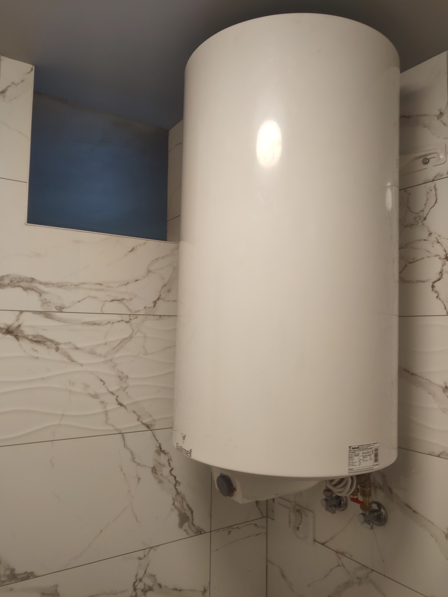 Bojler elektryczny Galmet zamontowany w łazience z marmurowymi płytkami, widoczne przyłącza wodne i elektryczne.