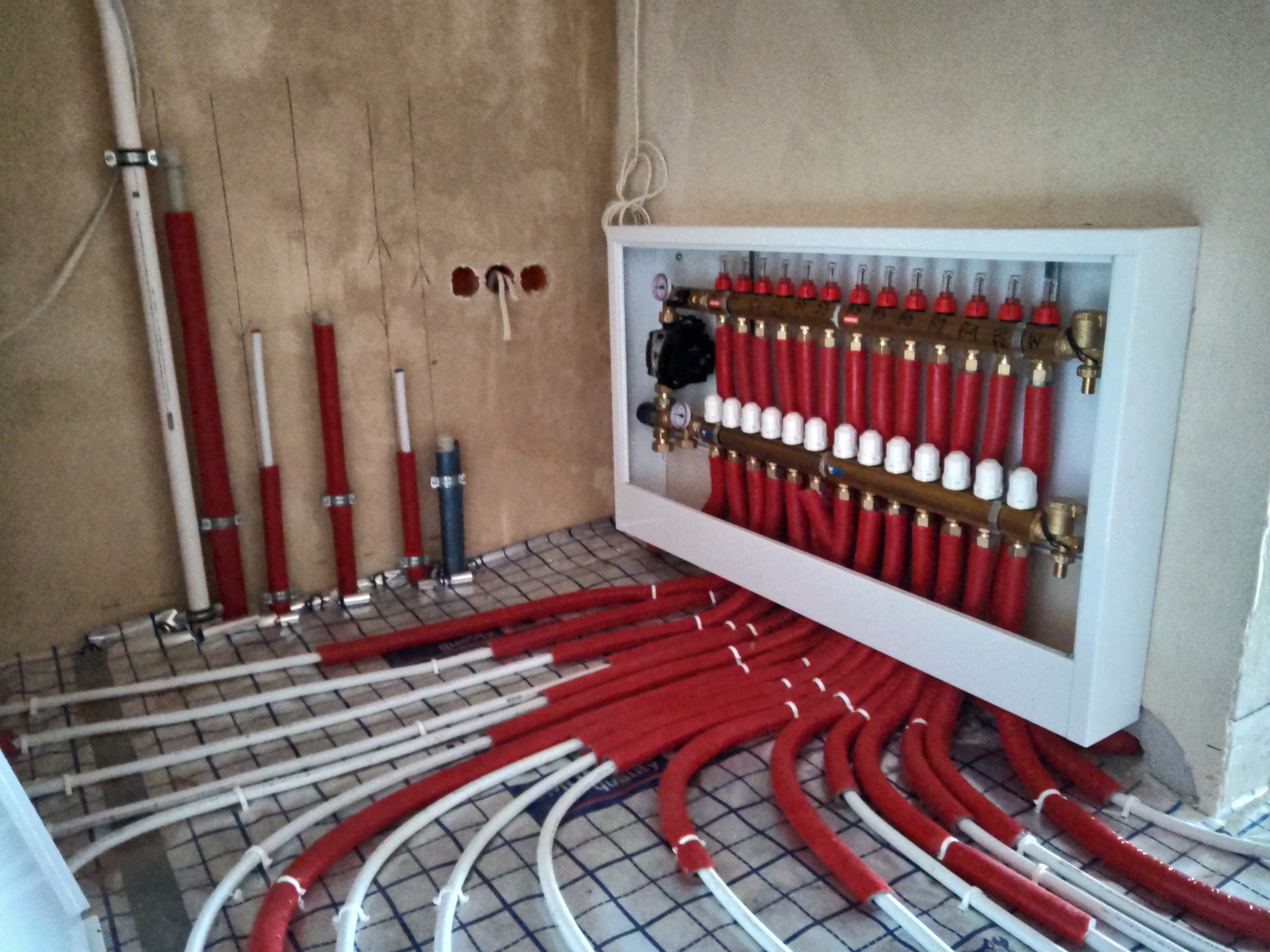 Rozdzielacz ogrzewania podłogowego z czerwonymi rurami na izolacji systemowej z widocznymi przyłączami do ściany w surowym stanie.