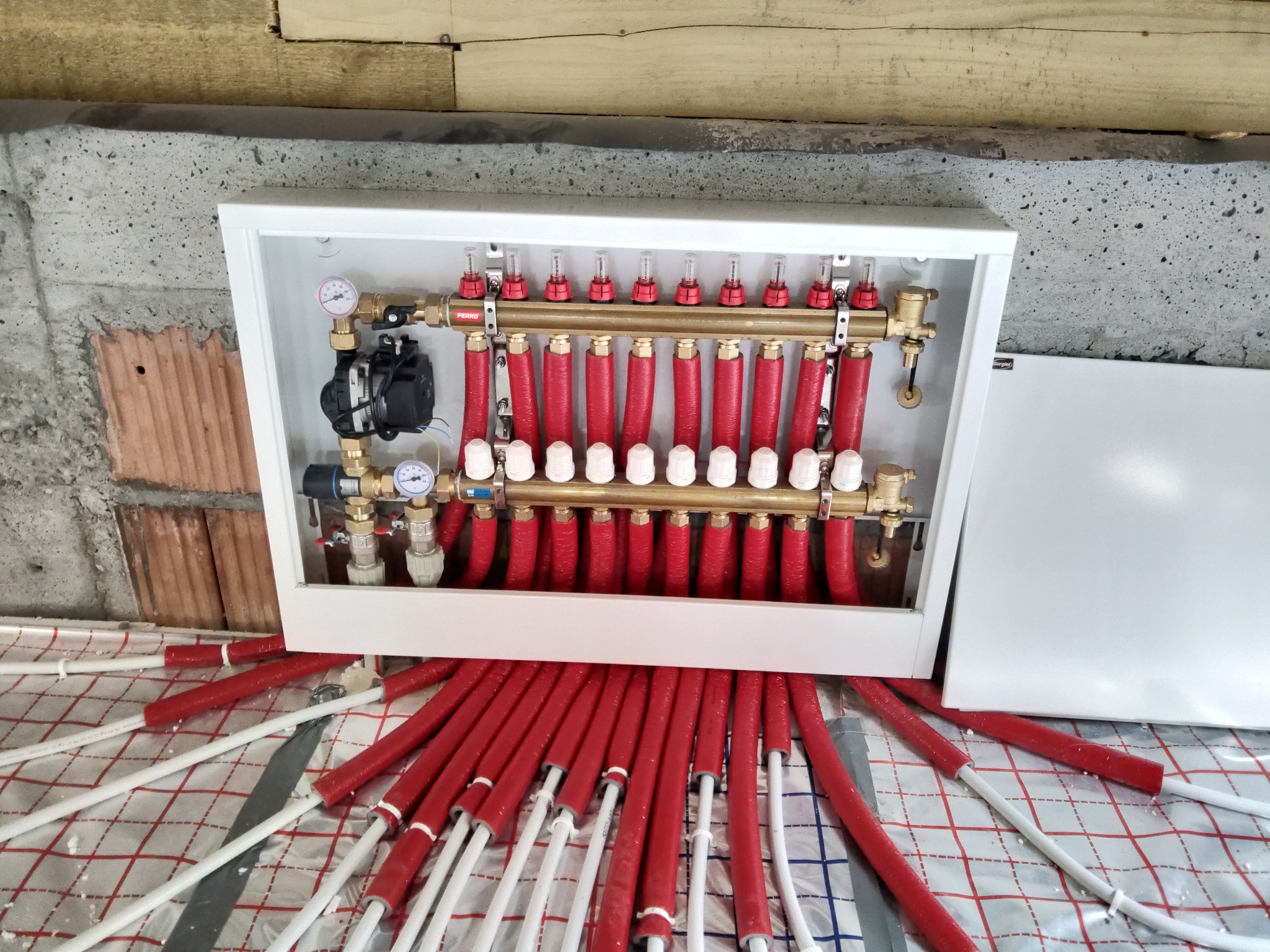 Rozdzielacz ogrzewania podłogowego w białej szafce, z widocznymi czerwonymi rurami grzewczymi na folii izolacyjnej z nadrukowaną siatką.