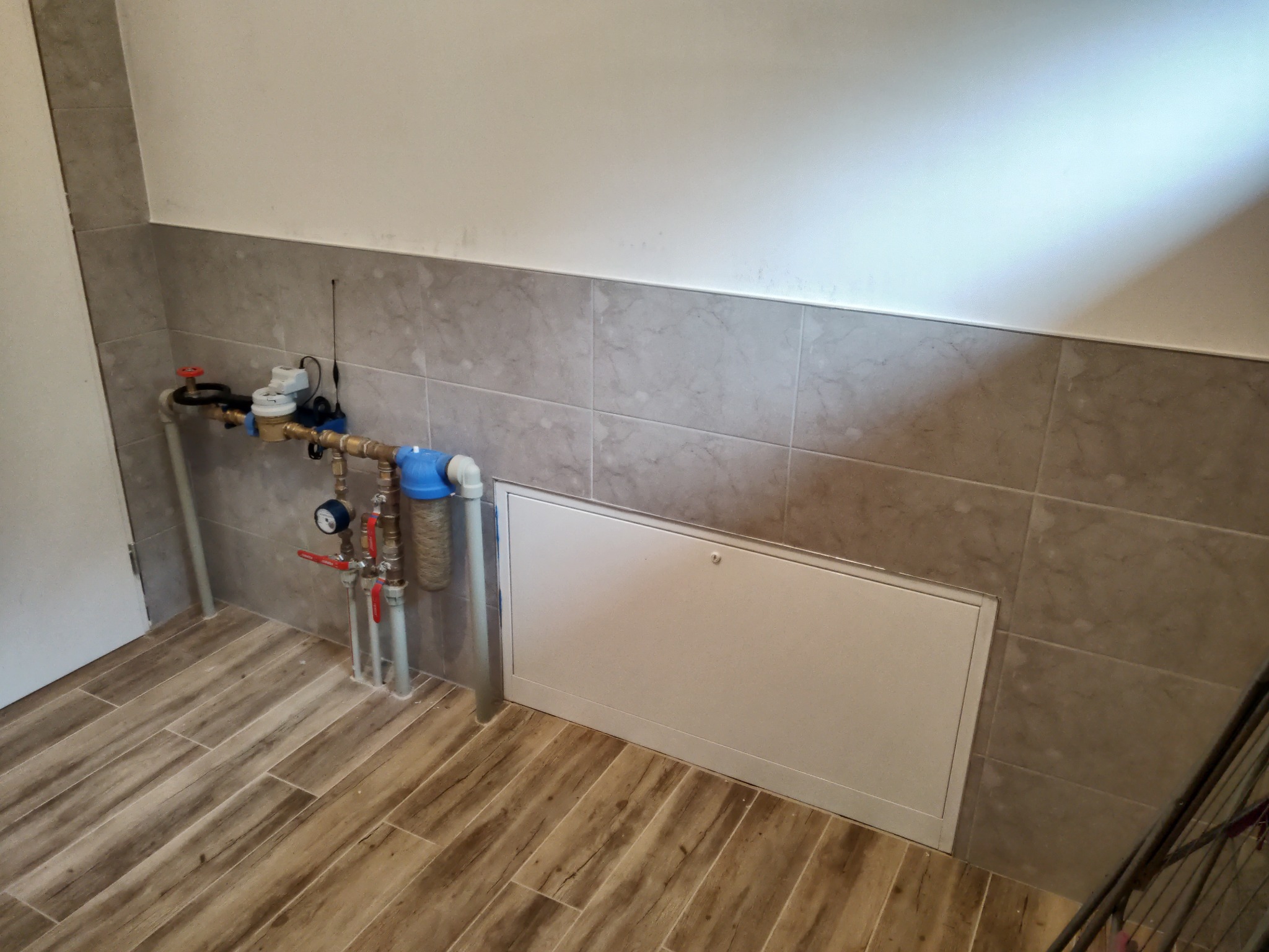 Węzeł wodomierzowy z filtrem narurowym i zaworami odcinającymi, zamontowany przy szarej ścianie z płytek i drewnopodobnej podłodze, z dostępem przez białą klapę rewizyjną.