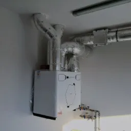 Biała pompa ciepła zamontowana na ścianie, z widocznymi, zaizolowanymi rurami wentylacyjnymi. Kompaktowa instalacja w jasnym pomieszczeniu.